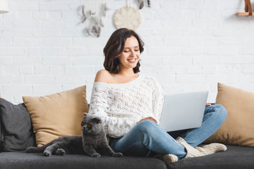 smiling girl using laptop with scottish fold cat on sofa