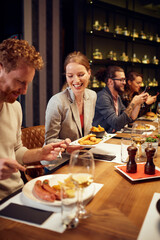 Handsome caucasian ginger taking food out of his girlfriend's plate while sitting in restaurant for dinner. In background are their friends.