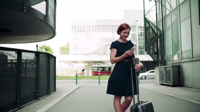 Young woman commuter with smartphone and suitcase standing outdoors in city.