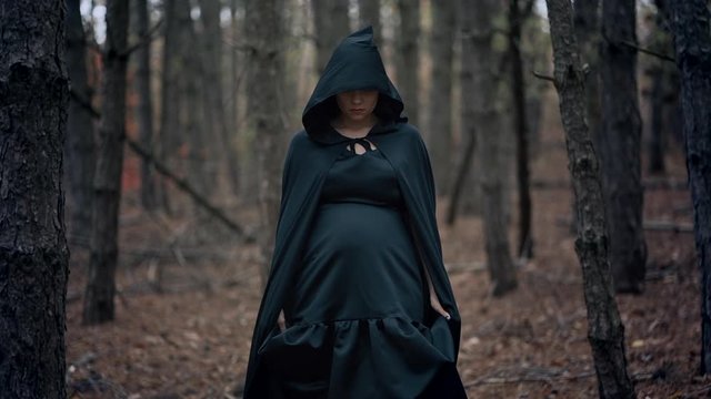 Black witch in hood walks in autumn scary forest.Womanl in long dress. Halloween, horror, concept, cosplay dressing up