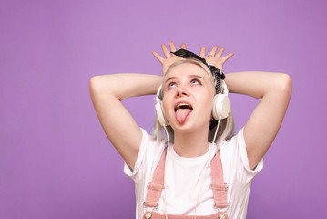 Cheerful girl in light clothing and headphones makes a funny face on a purple background. An emotional teen girl listening to music on a pink background.