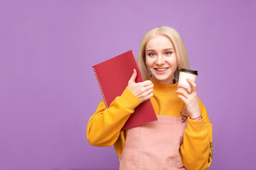 Happy blonde girl holds a cup of coffee and a notebook in her hands, looks at the camera and smiles, isolated on a purple background. Smiling student girl stands on a blue background and rejoices.