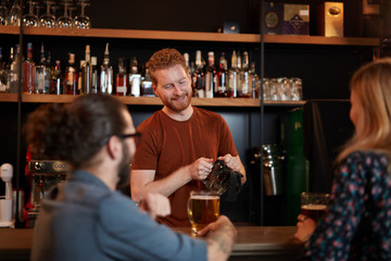 Handsome caucasian barman staning in bar and wiping pint and talking to customers. Customers drinking beer. Nightlife.