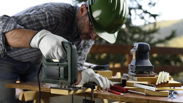 Adult carpenter craftsman wearing helmet and leather protective gloves, with electric saw working on cutting a wooden table. Construction industry, housework do it yourself. Footage.