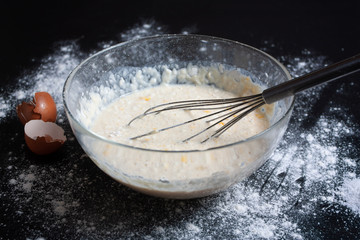 On a dark background is a glass bowl with ingredients for pancakes, eggs, flour, salt, milk. Traditional cuisine. Tasty food. Pancake Recipe. Dark background.