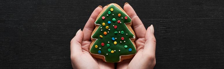 cropped view of woman holding Christmas tree cookie in hands on black wooden table, panoramic shot