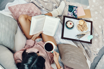 Top view of beautiful young woman in pajamas enjoying morning coffee while resting in bed at home