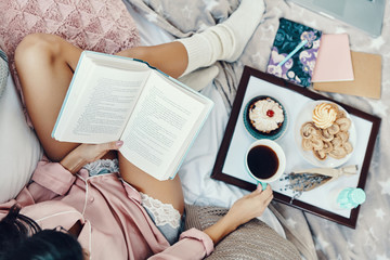  Top view of beautiful young woman in pajamas reading book and enjoying morning coffee while resting in bed at home