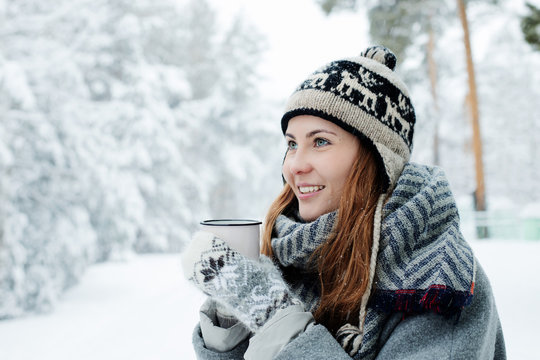 Beautiful young woman standing among snowy trees in winter forest and enjoying cup of hot tea.