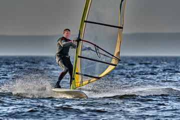 A male athlete is interested in windsurfing. He moves on a Sailboard on a large lake on an autumn day.