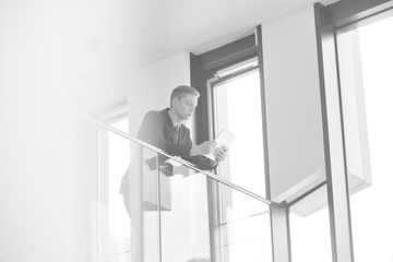 Black and white photo of Young businessman using digital tablet while leaning on railing at office