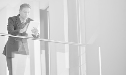Fototapeta premium Black and white photo of Young businessman using digital tablet while leaning on railing at office