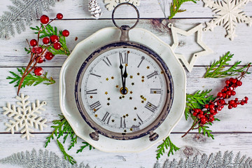 Christmas clock shows midnight. Antique clock with roman numerals on a white plate on a wooden light table near fir branches, cones, red berries, white snowflakes. Christmas and new year concept.