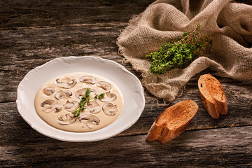 Plate with mushroom cream soup, pieces of toasted bread and a bundle of fresh thyme on sackcloth shred on wooden table. Low fat food concept. Warm colors