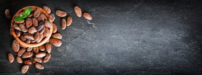 Cocoa beans on dark stone table top view, black background. Cocoas bean in wooden bowl with mint,...
