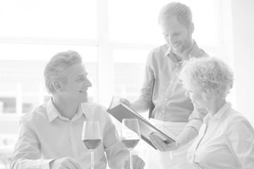Black and white photo of Young waiter showing menu to mature couple at restaurant