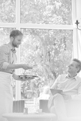 Young waiter serving coffee and food while mature customer using digital tablet at restaurant