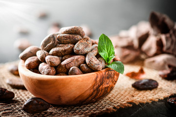 Cocoa beans with mint on jute side view. Cacao powder in wooden bowl blure in background. Panorama or banner concept.