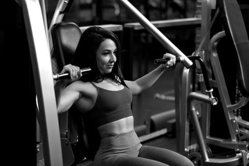 Fototapeta na wymiar Sexy fitness brunette girl is sitting on training apparatus and posing BW