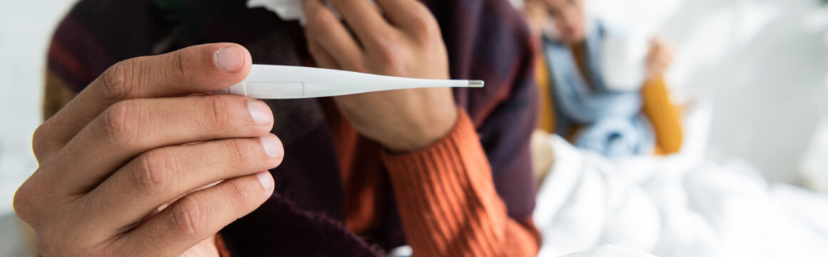 Cropped View Of Sick Man With Fever Holding Thermometer In Bedroom With Woman Behind