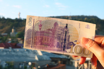 The Reverse Side of Georgian 100 Lari banknote in Woman's Hand with Blurry Tbilisi City View in the Backdrop