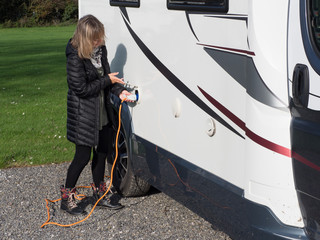 A lady standing lifts a flap and plugs in an orange electric hook up lead into the side of her motorhome to provide power to the camper van.Image