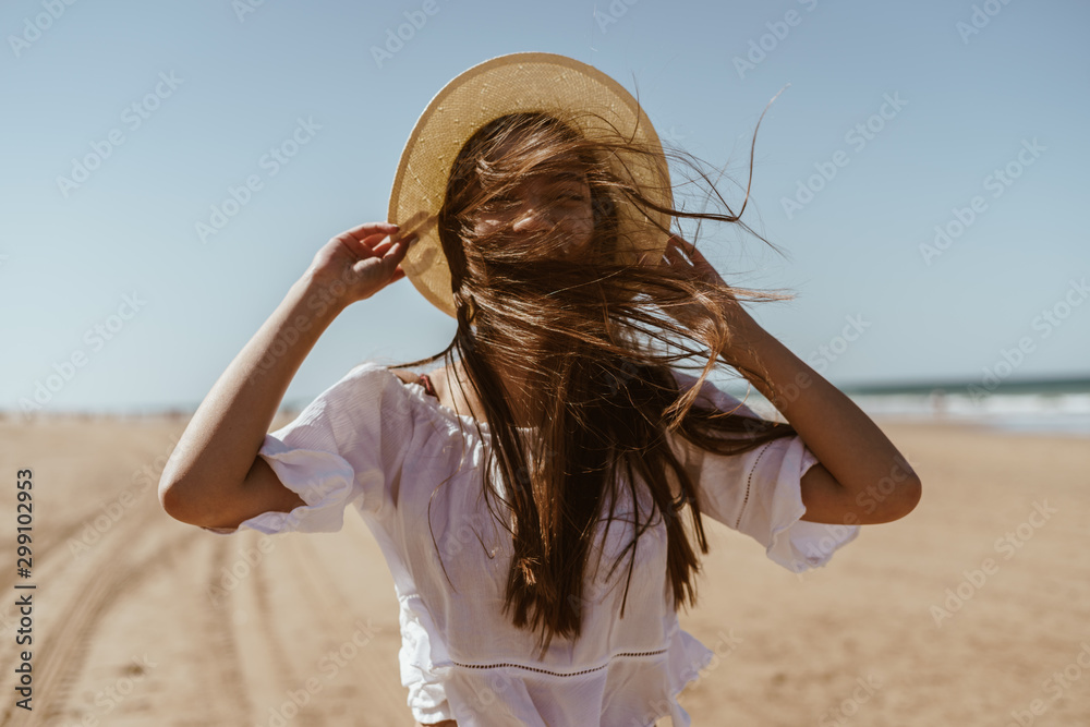 Wall mural Beach. The wind covered her face with her hair. She holds the hat with her hands.