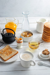 Healthy breakfast eating concept, various morning food - pancakes, soft-boiled egg, toast, oatmeal, granola, fruit, coffee, tea, orange juice, milk on white wooden table
