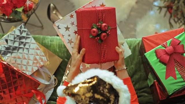 Holiday season. Upper view of stylish woman in sparkle gold sequin santa hat sitting on divan in the modern living room at Christmas taking Christmas present box from shopping bag. PRORes 422 codec.