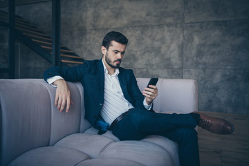 Portrait of his he nice good-looking imposing attractive luxury focused bearded guy wearing jacket sitting on divan using digital device in industrial interior room indoors