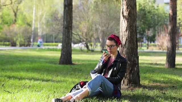 Beautiful young stylish woman sitting under a tree in the park with a cup of coffee and a telephone. girl making selfie in the park, sitting on the grass. stylish brunette in glasses with a bandage on
