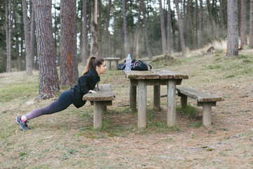 Fototapeta premium Fitness outdoor workout. Sporty young woman doing push ups using a rustic wood bench in the forest.