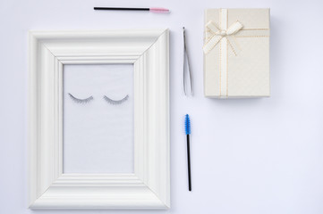 Beautiful eyelashes and eyebrows concept: white photo frame decorated with eyelash extensions and tools and gift on white background.