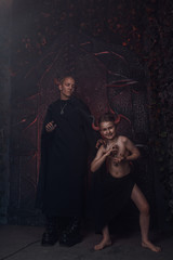 Naklejka premium small child with red horns of demon and adult scary man standing in front of the gates of hell. Halloween costumes of the father and the son ready for home party.