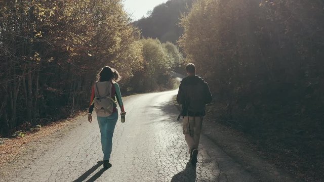 Hikers walking on a mountain road at sunrise from the back. Dolly shot of young man and woman trekkers walking through a forest.