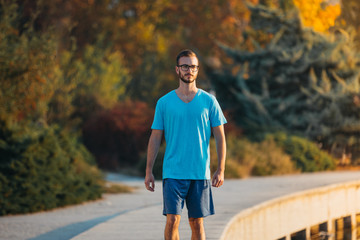 Obraz premium Young man with glasses in sportswear jogging by the lake in a city park. Runner training in the park by the water at sunset.