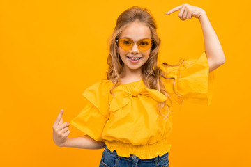 baby on a yellow background in a yellow dress and glasses shows gestures to herself