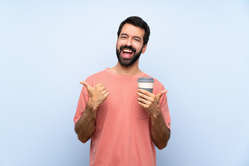 Young man with beard holding a take away coffee over isolated blue background with thumbs up gesture and smiling