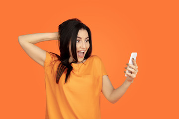 Caucasian young woman's portrait on orange studio background. Beautiful female brunette model in shirt. Concept of human emotions, facial expression, sales, ad. Copyspace. Making selfie, win in bet.