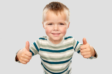 childhood, gesture and people concept - portrait of smiling little boy in striped shirt showing thumbs up over grey background
