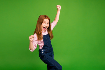 Profile photo of little ginger schoolchild celebrating winning classmates team in sportive competitions raise fists wear denim overall pink shirt isolated green background