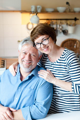 Happy senior couple is hugging and smiling in cozy home kitchen inside. Portrait of middle aged man and woman. Concept of wellbeing, happiness, kindness, male and female health, enjoying retirement.