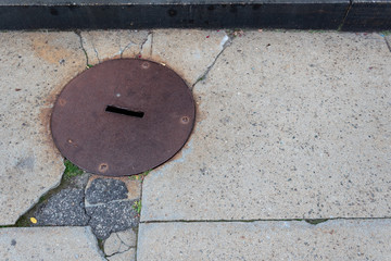 Rusted metal round manhole cover set in cracked sidewalk of concrete and asphalt, creative copy space, horizontal aspect
