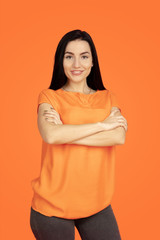 Obraz premium Caucasian young woman's portrait on orange studio background. Beautiful female brunette model in shirt. Concept of human emotions, facial expression, sales, ad. Copyspace. Standing hand crossed.