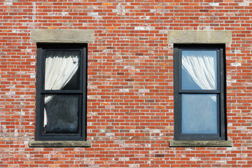 Two windows in a red brick wall. Window ledges are concrete, frames are wooden, painted black. Windows have white curtains, pulled right in one window and left in the other.