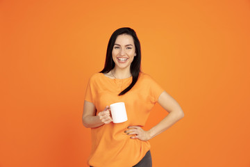 Caucasian young woman's portrait on orange studio background. Beautiful female brunette model in shirt. Concept of human emotions, facial expression, sales, ad. Copyspace. Drinking coffee or tea.