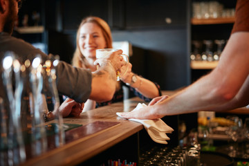 Bartender wiping bar counter while couple leaning on counter and making a toast with beer. Selective focus on hand.