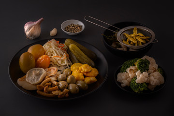 Fermented foods cabbage, peppers, pickles, tomatoes, mushrooms, zucchini, garlic in a large wooden dish on a dark table with spices for salting. Low key.