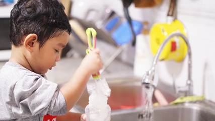 Two-year-old of Asian boy stands to wash his bottle in the kitchen alone. kid or baby help hose work on holiday concept.