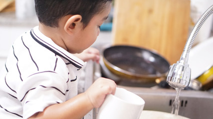 Two-year-old of Asian boy stands to wash his bottle in the kitchen alone. kid or baby help hose work on holiday concept.
